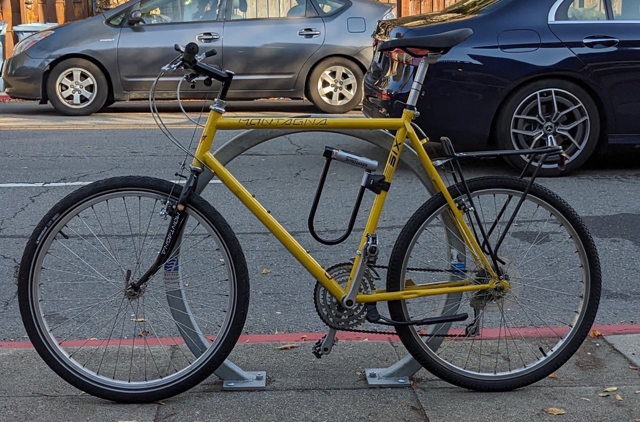 A yellow montagna six mountain bike locked to a rack on a street with cars