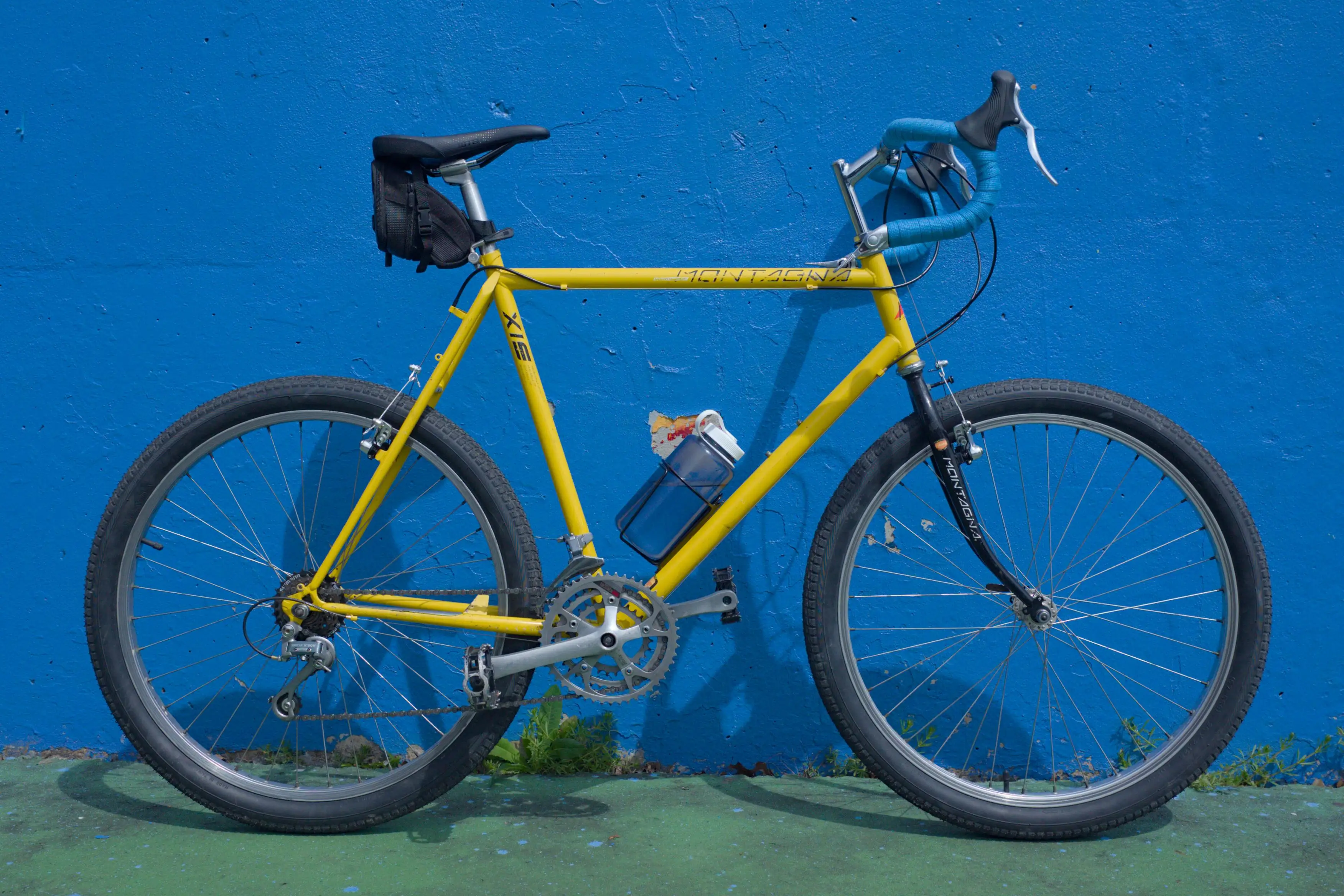 A yellow bike with blue handle bars in front of a blue wall.