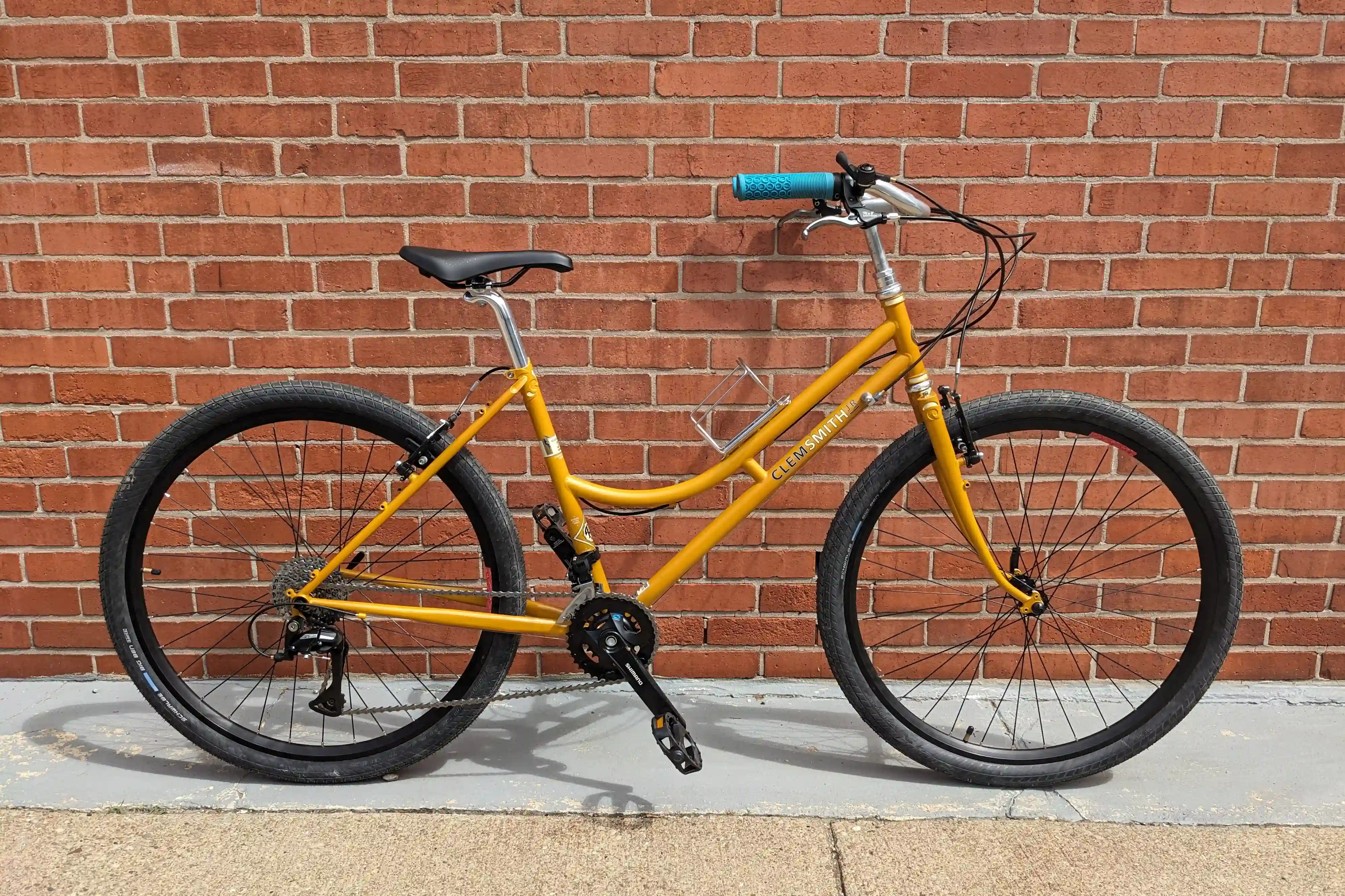 A gold bike leaning against a red brick wall. It has blue handle bar grips, mostly black components and a few silver accents