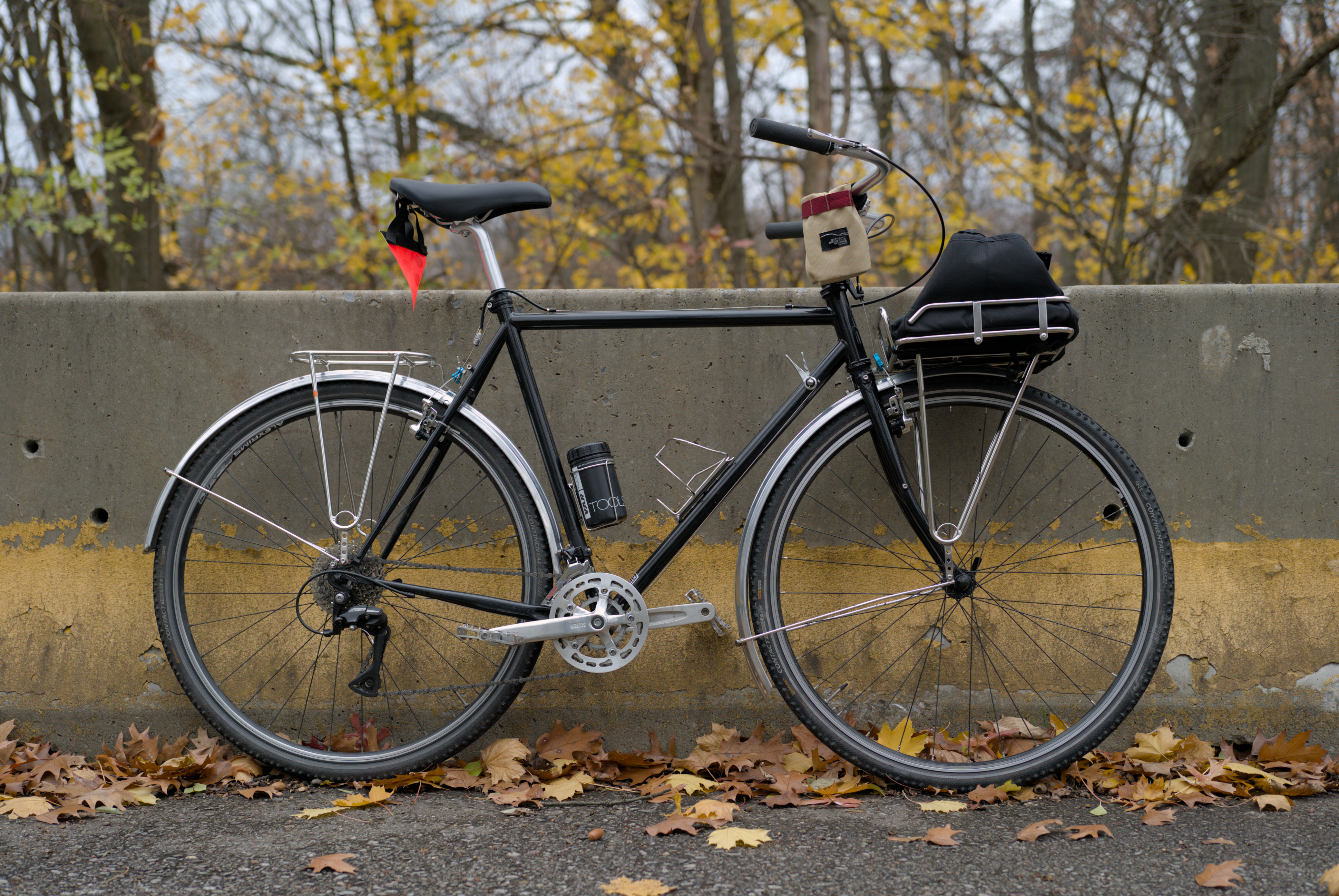 A black bike leaning against a concerete barrier. It has silver fenders, pedals, handle bars, and racks in the front and rear.